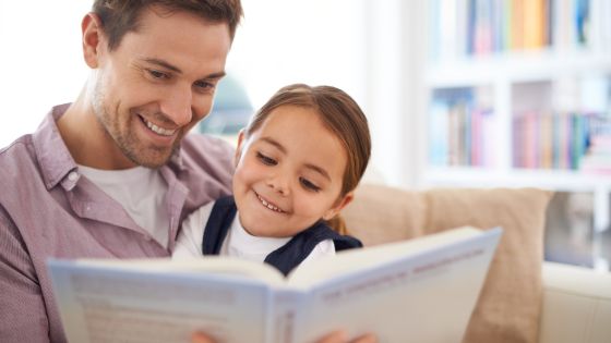 picture of a father smiling with his young daughter on his lap looking at a book together. In the background is a bookcase full of books.