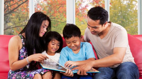 picture of a family with a mum, younger daughter, older son, and father all looking at a book together sat on the sofa