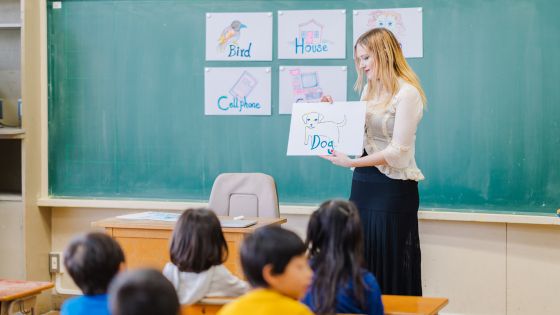6 children are sitting at their desks in a classroom looking at their female teacher. She has pictures and words on the blackboard of a bird, house, cell phone, and dog. It appears the children are learning English.