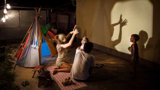 A family of four are sat outside, perhaps in the yard of their house. They are sat next to a teepee and in front of a light. They are making shadow pictures with their hands on the wall.