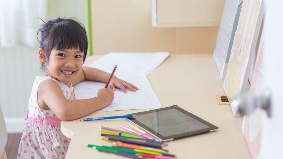 A girl is smiling sat at her desk with crayons on the table, an ipad and writing on a piece of paper.
