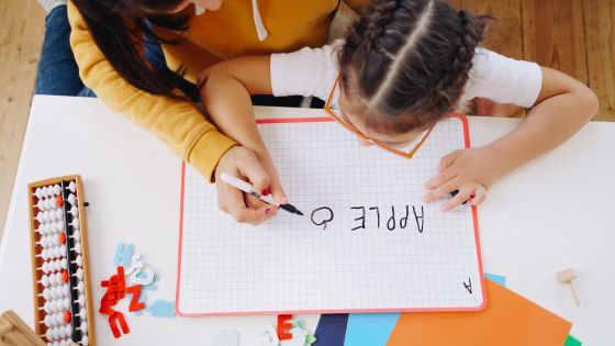 A picture of a girl being guided by either a teacher or guardian helping to write the word apple and draw a picture of an apple on a whiteboard.