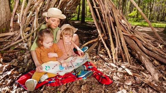 Three children sat together outside of a den made of sticks looking at a scavenger hunt map.