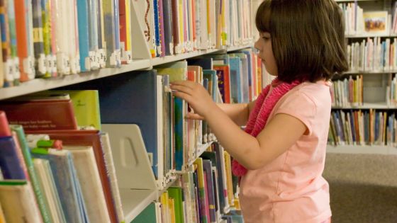 A young girl in a library looking through library books on the shelf.