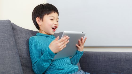 A boy sat on the sofa smiling at an ipad he is holding in his hands.