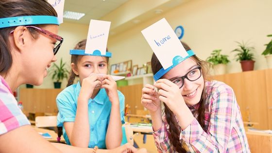 Three girls are sat together at a table laughing as they play a language game. They have bands on their head with a word written on a card for others to see. 