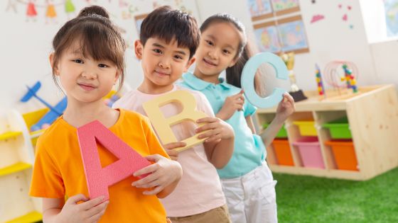 Three children looking at a the camera holding a big letter each "A B and C". They appear to be in kindergarten or preschool learning English.