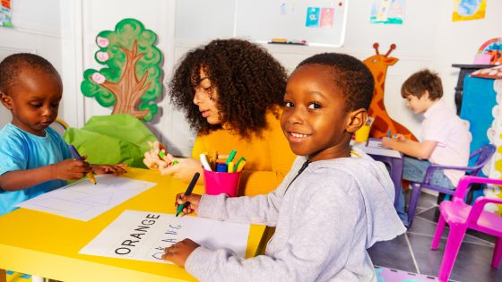Kindergarten class of 3-6 year olds learning English with a boy sat at the table writing out the word orange.