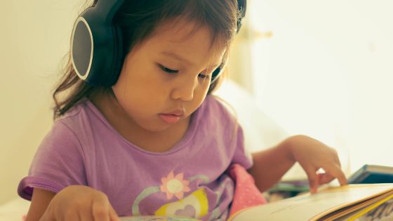 A girl sat down wearing large black headphones whilst looking at a book, possible listening to the audio book version.