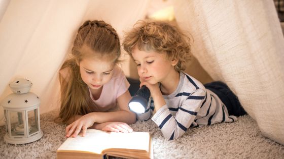Two children are lying on their fronts under a fabric tent with a torch light reading a fairy tale book together.