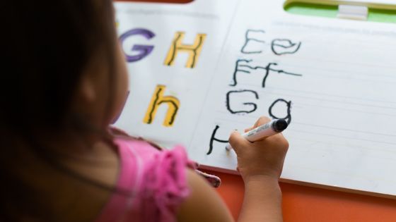 A girl writing at a table practicing English handwriting with the letters E, F, G, and H.