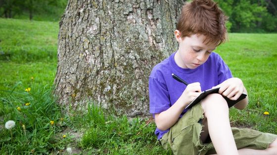 A boy sat by a tree writing in a journal.