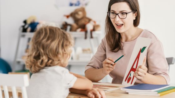A mum or teacher facing a boy sat at a desk holding up a picture of the letter A with an apple.