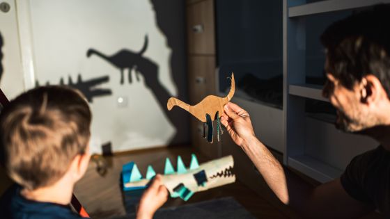 A male, possibly a dad, and a boy, possibly his son, with paper crafted dinosaurs in their hands. They are holding them in front of a light to make shadow puppets on the wall.