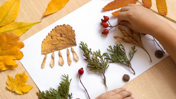Picture of a child's hand on a table making a craft on paper from natural objects e.g. leaves, small branches, and berries.