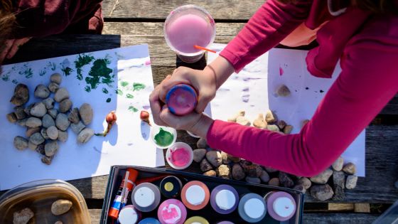 Children are sat outside on some wood with lots of round rocks and paint. They will be painting stones.
