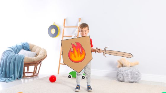 A boy is stood smiling with a homemade cardboard sword and shield that has a flame coloured on it.