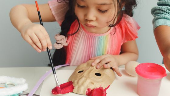 A girl is sat at a table painting a mask.