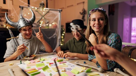 3 friends are playing a role-play board game together whilst wearing costumes e.g. a flower headband, Viking helmet and sword, a magicians hat.
