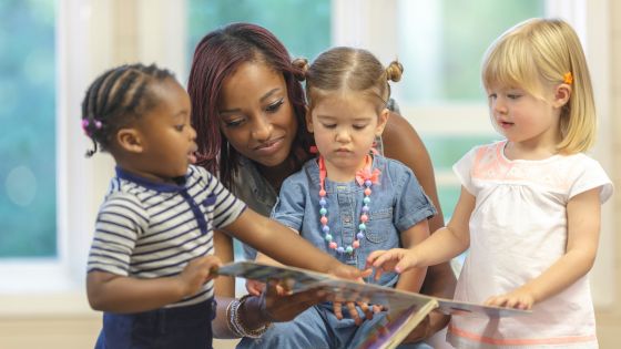 3 preschool-aged children are standing and sitting with a female school teacher looking at a book together.
