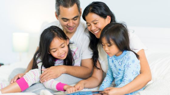 A family of four, mum, dad, and 2 daughters, are smiling whilst sitting together looking at a book