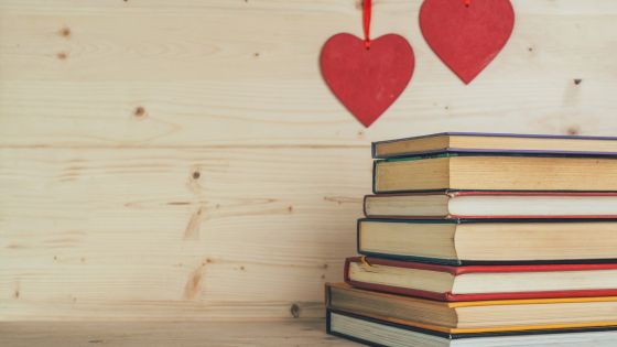 On a wooden table or bench, there are 7 books stacked on top of one another with two red plastic hearts hanging down over the top of them.