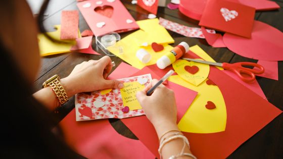 A table full of yellow, pink, and red coloured paper with glue and scissors. There are valentine's day cards on the table and a female is writing a valentine's day note inside one of the cards.