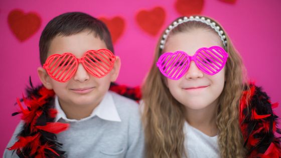 A boy and a girl wearing heart-shaped glasses with a pink background and paper hearts in the background.