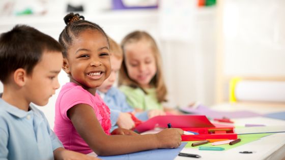 Image shows four children sat at a desk with crayons and paper. The girl in the middle is smiling and looking at the camera.