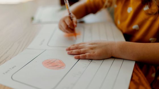 Image shows a child sat at a table with some worksheets and she is finishing a drawing on one of the worksheets.