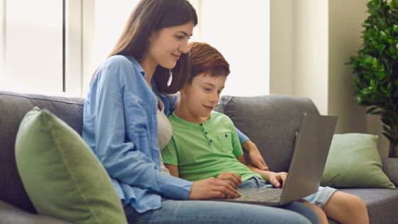 A mother and her son are sat together on the sofa looking at an open laptop.