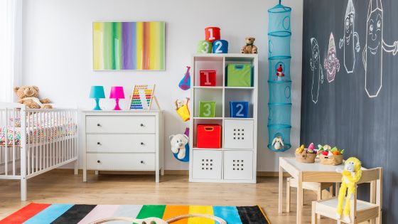 A colourful playroom with a cot, chest of draws, toy storage, desk and chairs, colourful rug, and a wall black board.