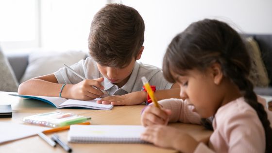 A boy and girl, perhaps brother and sister, are sat at a table together writing in books, maybe doing their homework.
