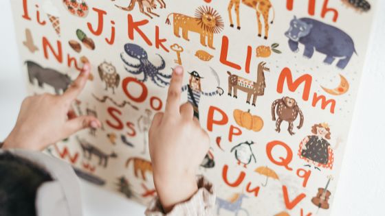 Image shows children's hands pointing to letters and images on an English alphabet poster.