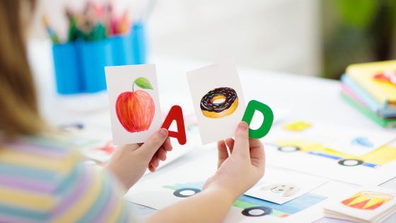 A girl is sat at a table with flash cards and she is holding an apple flash card and a donut flash card along with the letter's A and D.