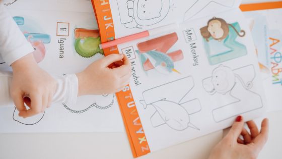 The image shoes an adult's hand and a child's hand looking at an open alphabet colouring book.