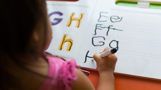 A young girl is sat at a table using whiteboard pen to trace over alphabet letters in a workbook.