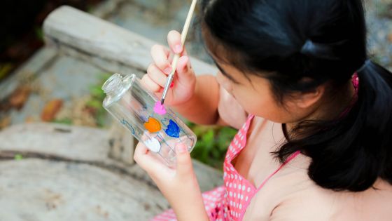 A girl is sitting outside by a wooden table. She is painting colourful hearts or flowers on a transparent bottle.
