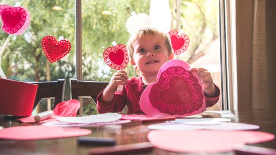 A young boy is sitting at a table holding up a pink and red paper heart and glue. It appears he is making a valentine's day card. There are other heart shaped papers on the table and heart shaped paper bunting on a window behind the boy.