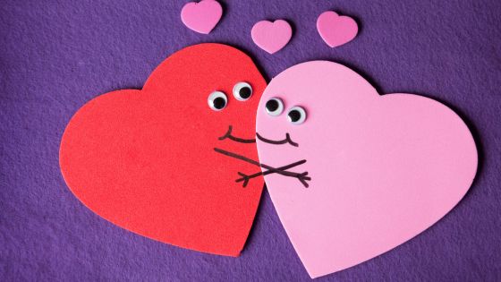 A picture of two felt hearts, one pink and one red, that have googly eyes attached and have pen drawn smiles and arms as if they are hugging one another. There are three small pink hearts in the background.