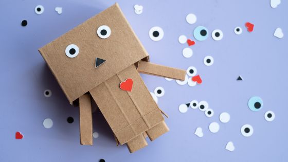 A cardboard robot with googly eyes, a triangle black nose, and a paper red heart. Behind, there are more heart shapes and googly eyes on the page.