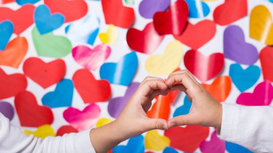 A wall covered in colourful paper hearts with two children's hands in the foreground joined together to form a heart shape.