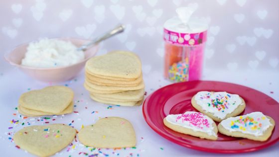 A picture with heart shaped biscuits, sprinkles, a bowl of white icing, and a red plate.