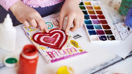 A girls' hands decorating a painted valentine's day card that has I love you mum written on it with a red painted heart.