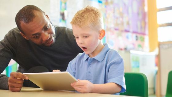 A picture of a man, perhaps a teacher, sat at a table with a school boy. They are holding a tablet and the boy is reading from it.
