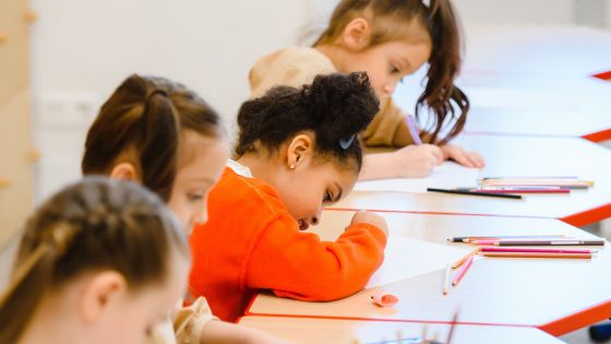 Four children sat in a row at desks, possibly in school, writing in their books.