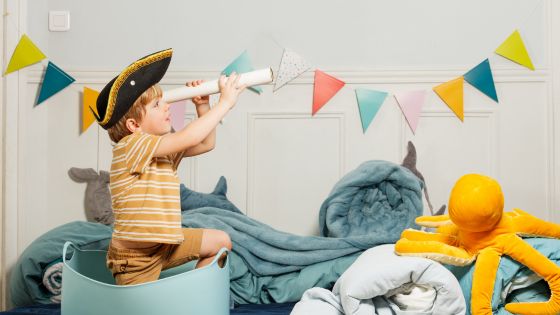 A boy playing make-believe as he stands in a basket pretending it is a boat. He has a pirate hat on and is looking out of a paper roll like a telescope. Behind him is blue bedding rolled like waves, a giant octopus teddy, and colourful bunting.