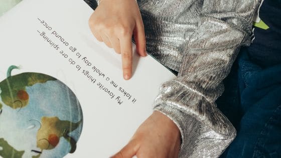 Picture shows a child sat down with an open book. The book is a picture book with an Earth illustration and the words "my favourite things to do are spinning it takes me a whole day to go around once."