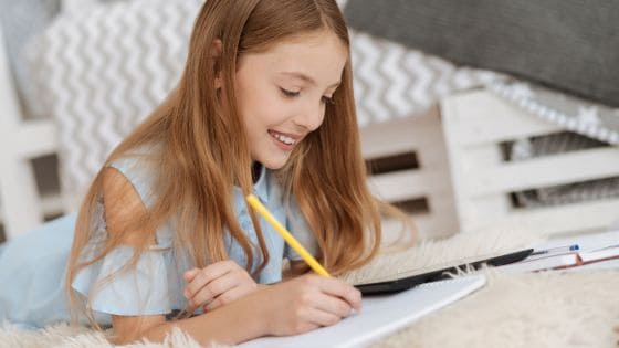 The image shows a girl in her bedroom lying down on the floor, smiling, as she writes on some paper.