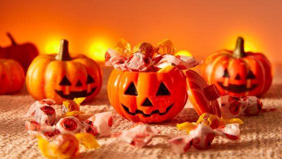 The photo shows a bright orange background with pumpkins. To the front are three tin jack-o-lanterns filled with candy/sweets. The centre tin has the lid off showing the candy inside and candy on the table.